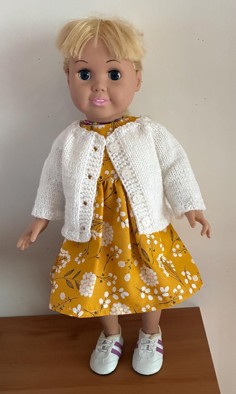 May include: A doll with blonde hair wearing a mustard yellow dress featuring a white floral pattern, a white knitted cardigan with four buttons, and white sneakers with purple stripes. The doll has blue eyes and pink lips.