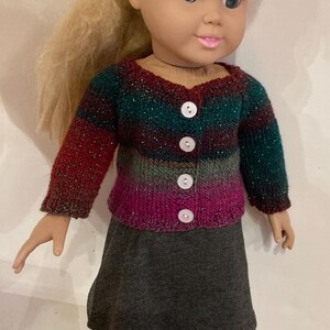 May include: A doll with blonde hair wearing a long-sleeved, multi-colored knit cardigan featuring horizontal stripes in shades of dark green, red, and magenta with subtle metallic flecks, fastened with four white buttons over a plain gray skirt.
