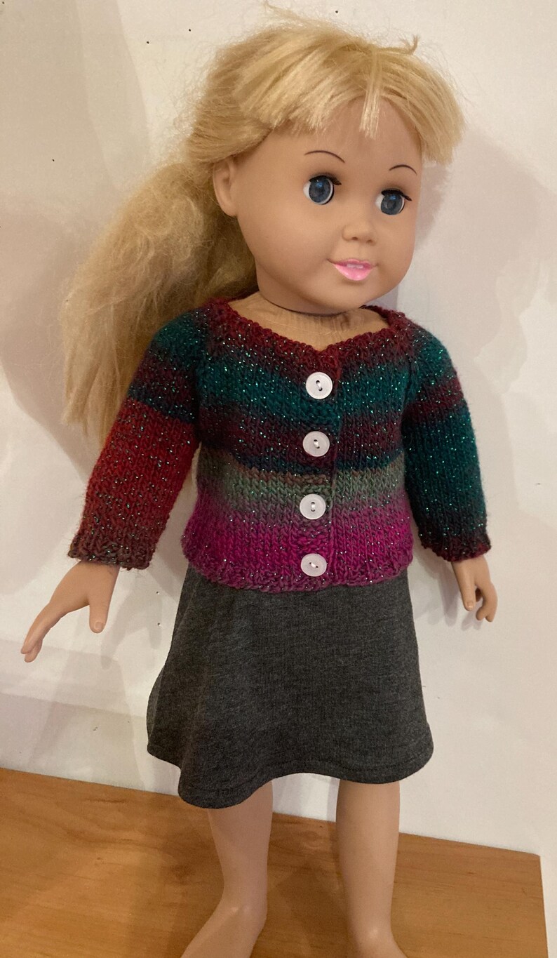 May include: A doll with blonde hair wearing a long-sleeved, multi-colored knit cardigan featuring horizontal stripes in shades of dark green, red, and magenta with subtle metallic flecks, fastened with four white buttons over a plain gray skirt.
