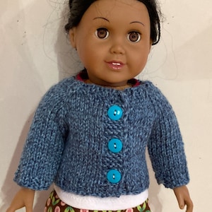 May include: A doll with dark brown hair and brown eyes wearing a blue knitted cardigan with three bright turquoise buttons. Underneath the cardigan is a white shirt and a brown skirt featuring a repeating pattern of green leaves and pink flowers.