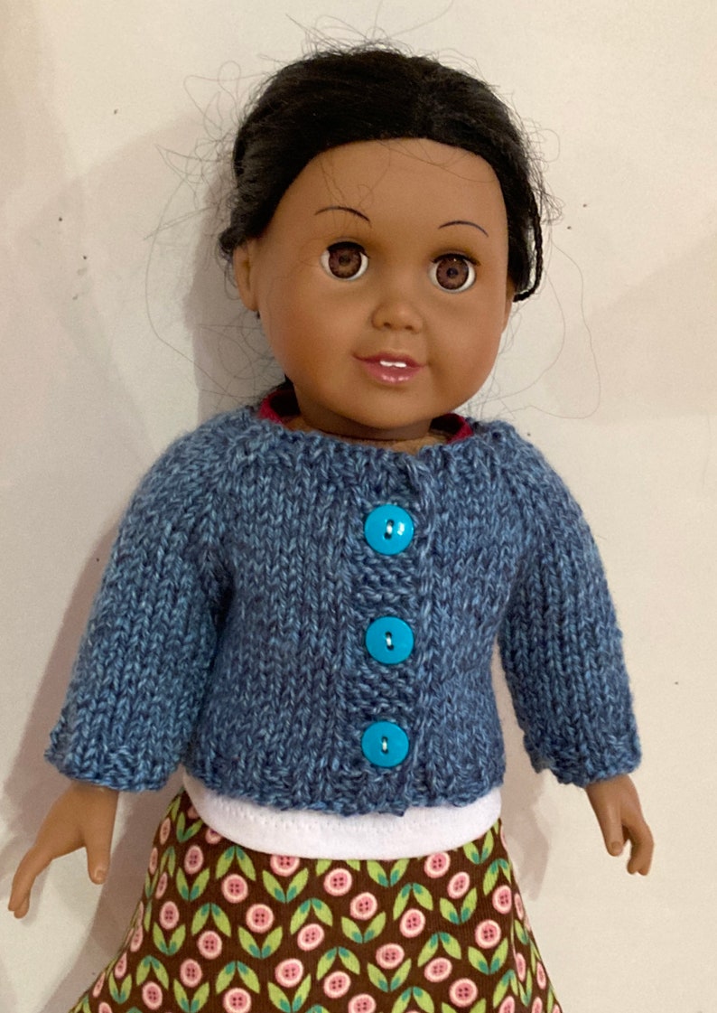 May include: A doll with dark brown hair and brown eyes wearing a blue knitted cardigan with three bright turquoise buttons. Underneath the cardigan is a white shirt and a brown skirt featuring a repeating pattern of green leaves and pink flowers.