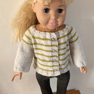 May include: A doll with blonde hair wearing a white and olive green striped knit cardigan with four clear buttons and dark gray leggings. The doll has blue eyes and pink lips, and the cardigan features a ribbed neckline and cuffs.