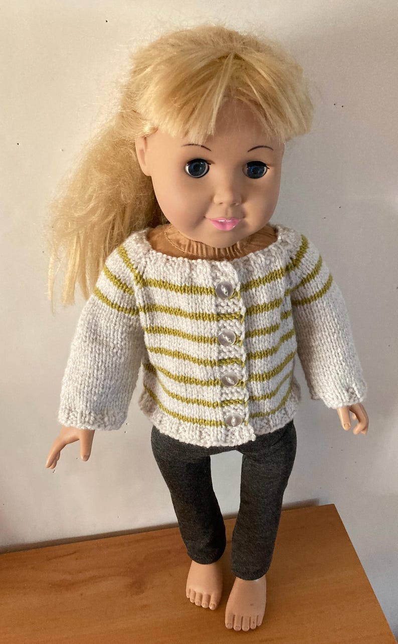 May include: A doll with blonde hair wearing a white and olive green striped knit cardigan with four clear buttons and dark gray leggings. The doll has blue eyes and pink lips, and the cardigan features a ribbed neckline and cuffs.