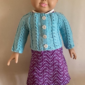 May include: A doll with blonde hair wearing a light blue knitted cardigan with four white buttons, a purple skirt featuring a white floral pattern, and white sneakers with purple accents. The doll has blue eyes and a neutral facial expression.