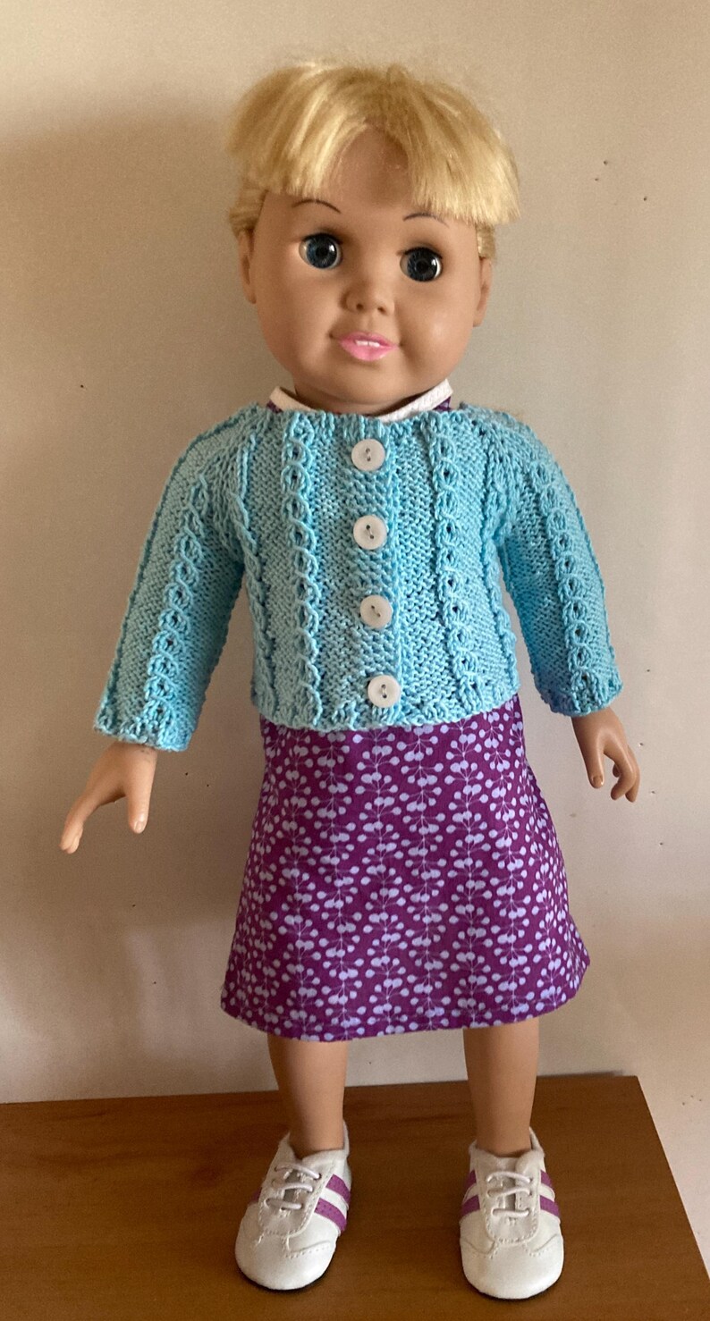 May include: A doll with blonde hair wearing a light blue knitted cardigan with four white buttons, a purple skirt featuring a white floral pattern, and white sneakers with purple accents. The doll has blue eyes and a neutral facial expression.