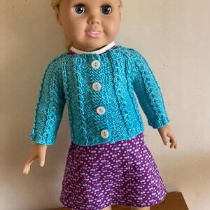 May include: A doll with blonde hair wearing a bright turquoise knitted cardigan with four white buttons over a purple dress featuring a white leaf pattern. The doll is also wearing white sneakers with purple stripes on the sides and a white collar.