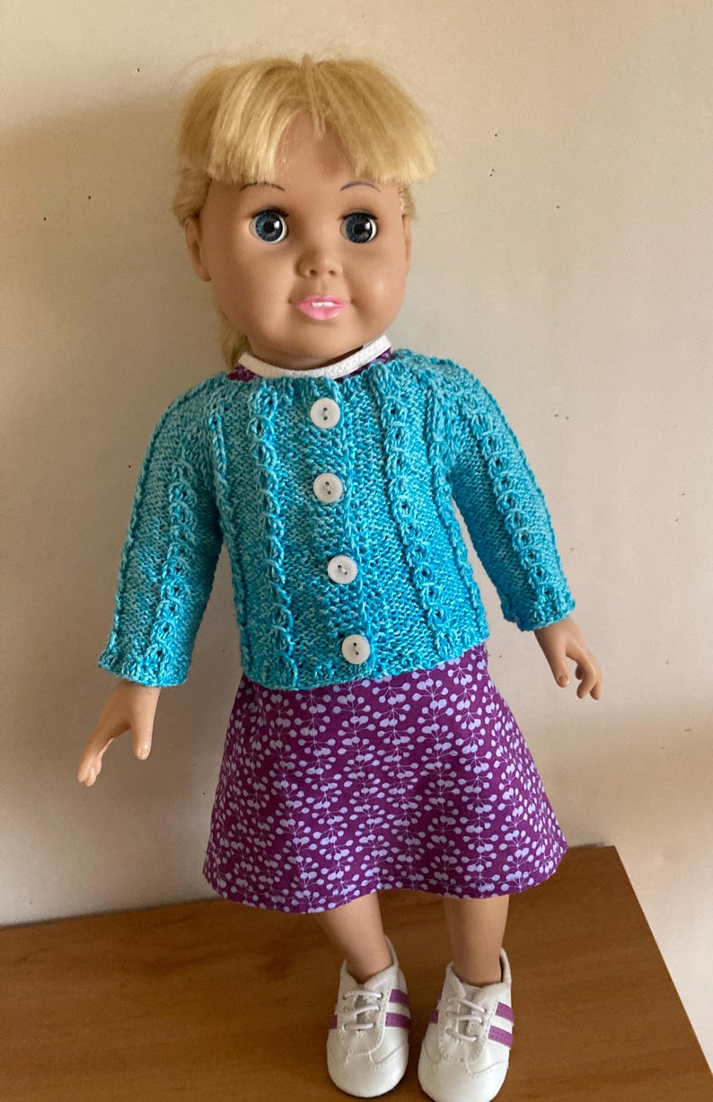 May include: A doll with blonde hair wearing a bright turquoise knitted cardigan with four white buttons over a purple dress featuring a white leaf pattern. The doll is also wearing white sneakers with purple stripes on the sides and a white collar.