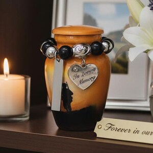 May include: An orange and black memorial urn with a decorative beaded bracelet and a heart-shaped charm that reads "In Loving Memory." A candle burns in a glass holder, and a framed picture and flowers are in the background.