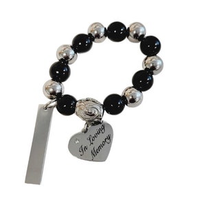 May include: A beaded bracelet with alternating black and silver-colored beads. A heart-shaped charm with the words "In Loving Memory" is attached, along with a rectangular tag. The bracelet is designed to be worn on the wrist.