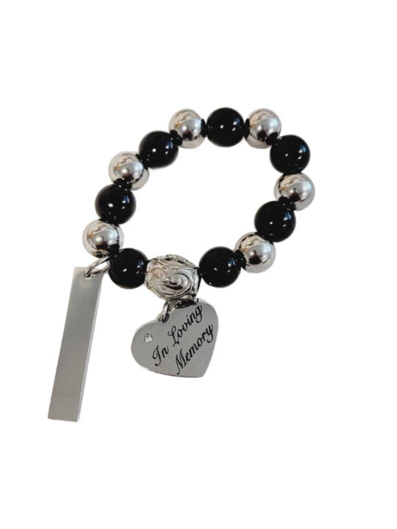 May include: A beaded bracelet with alternating black and silver-colored beads. A heart-shaped charm with the words "In Loving Memory" is attached, along with a rectangular tag. The bracelet is designed to be worn on the wrist.