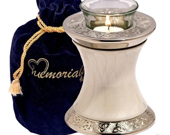 May include: White ceramic urn with a silver metal top and a glass candle holder. The urn has a Celtic knot design around the base and a textured pattern on the body. It comes with a blue velvet bag with a gold drawstring and the word "Memorial" embroidered on it.