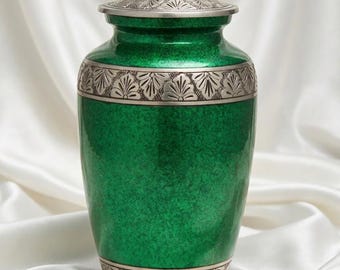 Puede incluir: Una urna funeraria de color verde metálico con un acabado moteado y bandas plateadas en la parte superior e inferior. Las bandas plateadas están decoradas con un patrón en relieve en forma de abanico. La urna tiene una tapa plateada a juego.