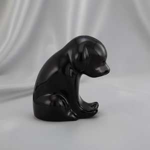 May include: A glossy black dog figurine in a seated pose. The sculpture has a smooth, reflective surface and a minimalist design. The dog's head is slightly lowered, and its paws are tucked beneath its body.