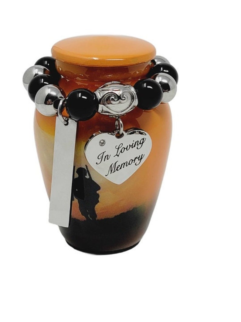 May include: An orange and brown cremation urn with a sunset scene and a soldier silhouette. It has a heart-shaped pendant that says "In Loving Memory". The urn is adorned with a beaded bracelet of black and silver beads.