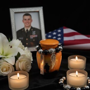 May include: An orange and black memorial urn with a silver charm and a beaded bracelet. A framed photo, flowers, candles, and an American flag are in the background. The urn has the words "In Loving Memory" on it.