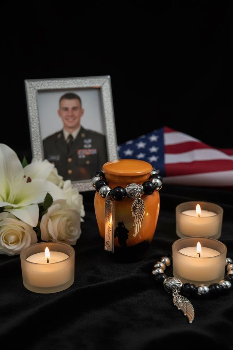 May include: An orange and black memorial urn with a silver charm and a beaded bracelet. A framed photo, flowers, candles, and an American flag are in the background. The urn has the words "In Loving Memory" on it.