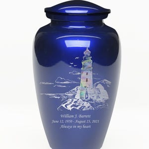 K&ouml;nnte beinhalten: Blaue Metall-Urne mit einem wei&szlig;en, gravierten Bild eines Leuchtturms an einer felsigen K&uuml;ste. Die Urne ist mit dem Namen "William J. Barrett", den Daten "12. Juni 1950 - 23. August 2021" und dem Satz "Immer in meinem Herzen" beschriftet.