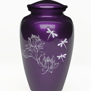 May include: A deep purple cremation urn with a glossy finish, featuring a silver-toned engraved design of two lotus flowers on the left and three dragonflies on the right. The vessel has a rounded body and a matching removable lid on top.