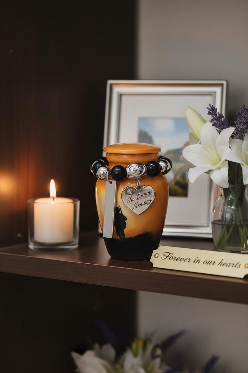 May include: An orange and black memorial urn with a decorative beaded bracelet and a heart-shaped charm that reads "In Loving Memory." A candle burns in a glass holder, and a framed picture and flowers are in the background.
