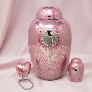 May include: Pink and silver urn set with a rose design. The set includes a large urn, a small keepsake urn, and a heart-shaped memorial. The urns have a decorative floral pattern.