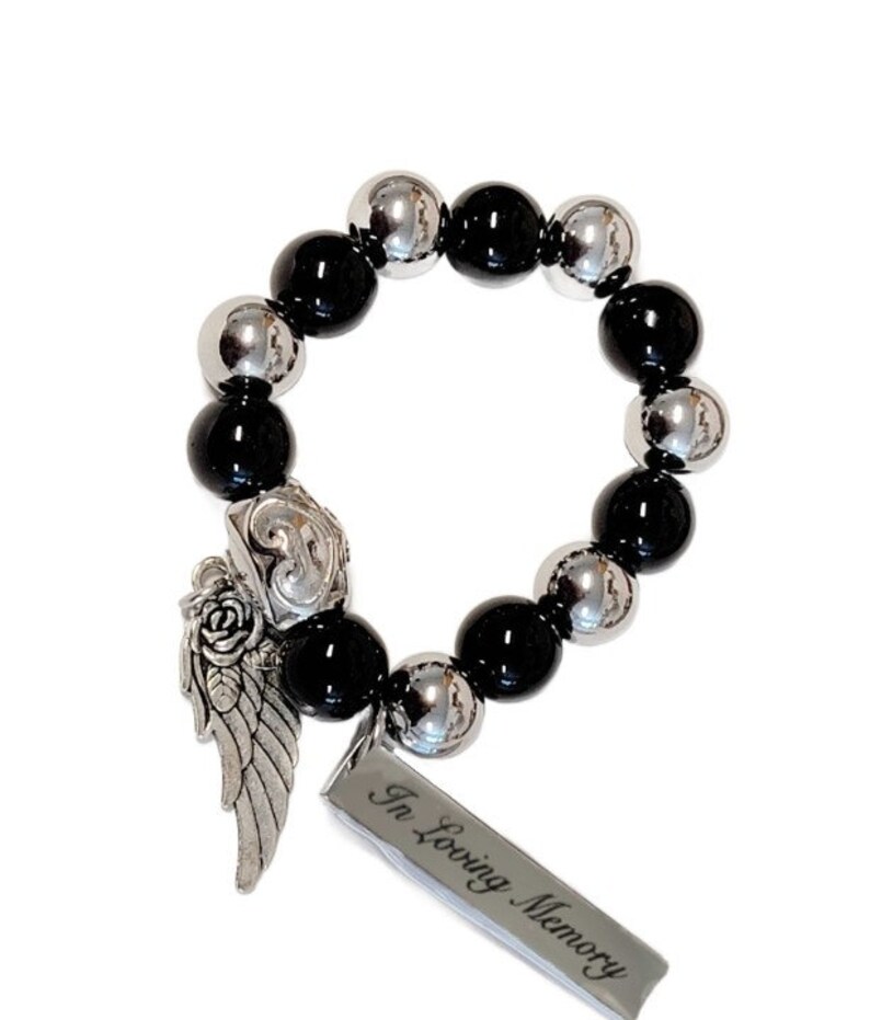 May include: A beaded bracelet featuring alternating black and silver-colored beads. A silver charm with an angel wing and rose design is attached, along with a rectangular tag that reads "In Loving Memory".
