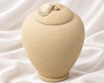 May include: A beige, textured, urn-shaped vessel with a matching lid. The lid is adorned with a single, light-colored, natural seashell centered on top. The container has a rounded body that tapers toward a circular base, featuring a matte finish.
