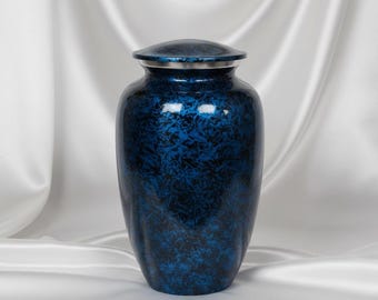 May include: A cylindrical metal cremation urn featuring a deep blue and black marbled finish. The vessel has a smooth, rounded body that tapers toward the base and includes a matching lid with a thin, polished silver-toned metal band at the rim.