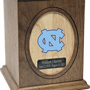 Puede incluir: Una urna de madera con un acabado marr&oacute;n claro. La parte delantera de la urna presenta una incrustaci&oacute;n circular con un fondo de madera clara y un logotipo azul de los North Carolina Tar Heels. Una placa plateada debajo del logotipo dice "William J Barrett 12 de junio de 1950 - 23 de agosto de 2021".