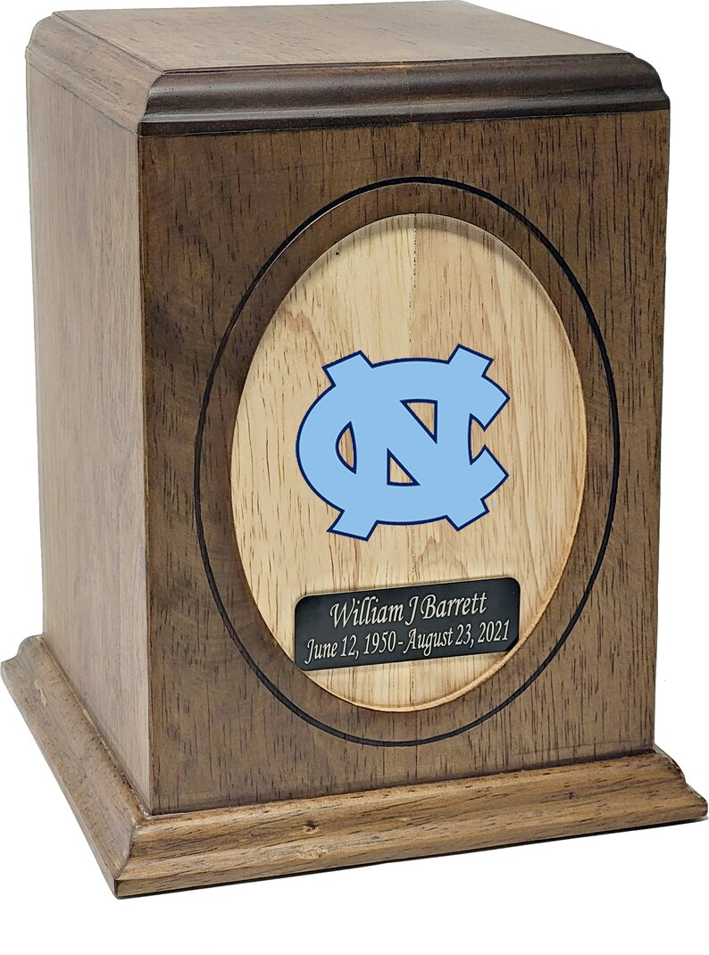 Puede incluir: Una urna de madera con un acabado marr&oacute;n claro. La parte delantera de la urna presenta una incrustaci&oacute;n circular con un fondo de madera clara y un logotipo azul de los North Carolina Tar Heels. Una placa plateada debajo del logotipo dice "William J Barrett 12 de junio de 1950 - 23 de agosto de 2021".