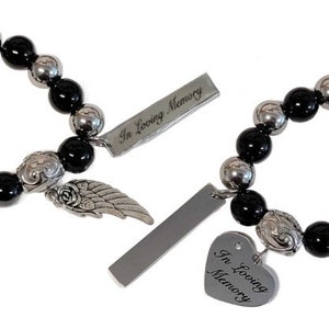 May include: Two beaded bracelets with black and silver beads. Each bracelet has a decorative charm with a rose and wing design, and a rectangular tag with the words "In Loving Memory". One bracelet has a heart-shaped tag.