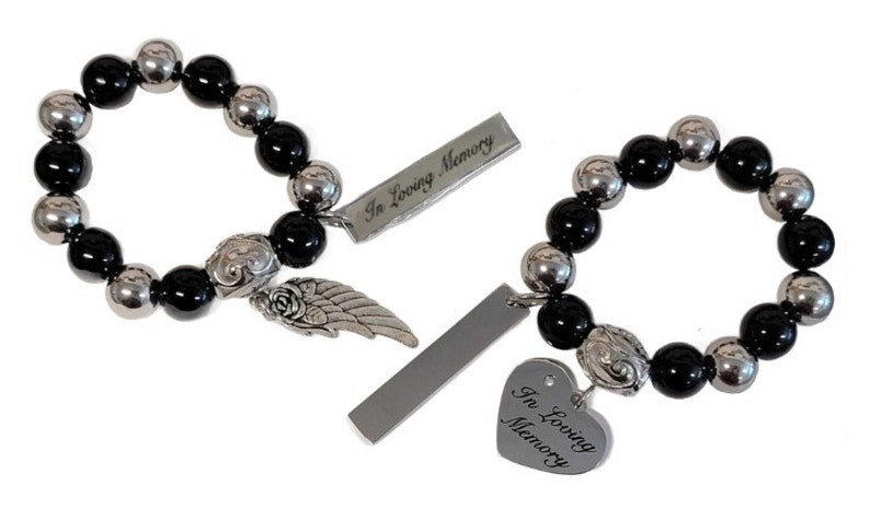 May include: Two beaded bracelets with black and silver beads. Each bracelet has a decorative charm with a rose and wing design, and a rectangular tag with the words "In Loving Memory". One bracelet has a heart-shaped tag.