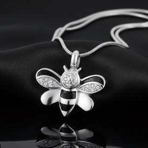 May include: A silver-colored bee pendant necklace. The bee pendant has a detailed design with clear crystals on the wings and body. The necklace chain is also silver-colored and has a delicate, snake-like design. The pendant is approximately 1 inch in length.
