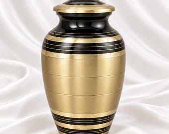 May include: A cylindrical cremation urn featuring a polished black finish on the lid and neck, contrasted by a brushed gold body. The design includes horizontal black bands and thin gold pinstripes encircling the upper and lower sections.
