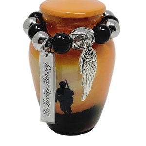 May include: An orange and black memorial urn with a sunset scene and a soldier silhouette. It features a beaded bracelet with black and silver beads, a silver charm with a rose and wing design, and a tag that reads "In Loving Memory."