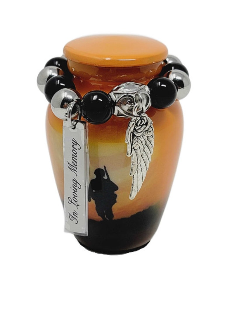 May include: An orange and black memorial urn with a sunset scene and a soldier silhouette. It features a beaded bracelet with black and silver beads, a silver charm with a rose and wing design, and a tag that reads "In Loving Memory."