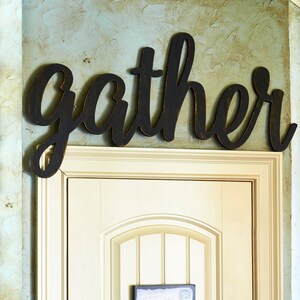 May include: Black wooden sign with the word "gather" in a cursive font. The sign is mounted above a white door with a framed print of a pig.
