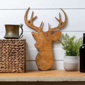 May include: A wooden cutout of a deer head with antlers, painted in a brown stain. The deer head is mounted on a white wall, with a woven basket and a copper mug in the foreground.
