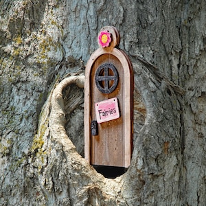 May include: A wooden fairy door with a pink flower design and a sign that reads "Fairies" is set into the bark of a tree.