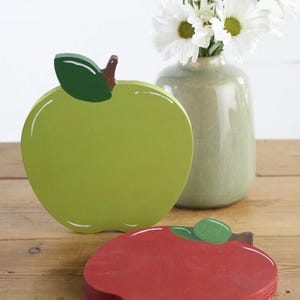 May include: Two wooden apples, one green and one red, with painted details. The green apple has a brown stem and a green leaf. The red apple has a green leaf. The apples are on a wooden surface.