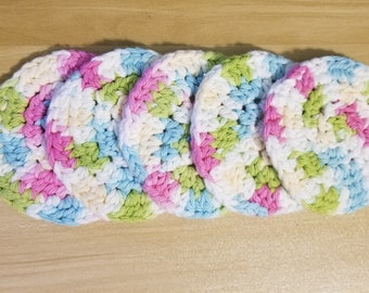 May include: Five crocheted coasters in a circular shape with a multi-coloured pattern of pink, blue, green, and white.