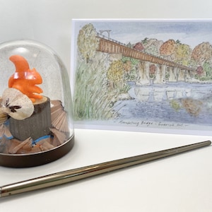 May include: A glass dome with a small orange squirrel figurine sitting on a wooden stump. The dome is filled with pencil shavings. A watercolor painting of a bridge over a river is in the background. The painting is titled "Monessen Bridge - Frederick, Md."