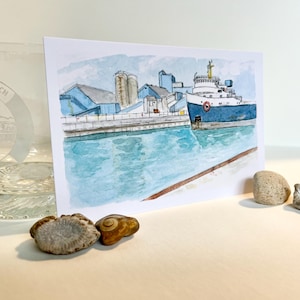 May include: A watercolour postcard depicting a blue cargo ship docked at the Port of Goderich, next to a clear glass etched with the same port name and image, accompanied by four small natural stones arranged on a white surface.