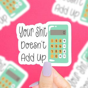 May include: A sticker with a calculator and the text "Your Shit Doesn't Add Up". The calculator is green and has a white display with the number 0 showing. The sticker is held by a hand with a light purple nail.