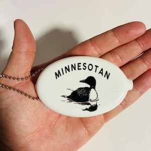 May include: White oval coin purse with a black and white illustration of a loon on water. The word "MINNESOTAN" is printed above the illustration in black. A silver ball chain is attached to the purse.
