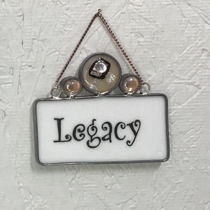 May include: White glass sign with a silver frame and the word "Legacy" written in black cursive. The sign is hanging from a brown chain with three decorative glass beads.