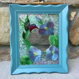 May include: A framed stained glass art piece featuring a hummingbird and flowers. The hummingbird has red and purple feathers, with a long beak. The flowers are blue and purple. The frame is a light blue color.