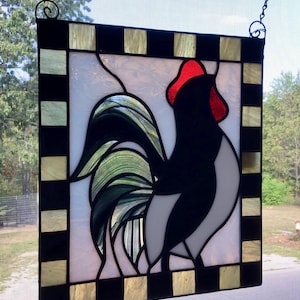 May include: Stained glass panel featuring a stylized rooster. The rooster is depicted in black, white, and green, with a red comb. The panel is framed with a black and gold checkered border and includes a chain for hanging.