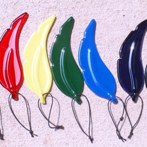Puede incluir: Un conjunto de ocho adornos de plumas de vidrio de colores. Las plumas son de varios colores, incluyendo rojo, blanco, amarillo, verde, azul y negro. Cada pluma tiene un lazo en la parte superior para colgar.