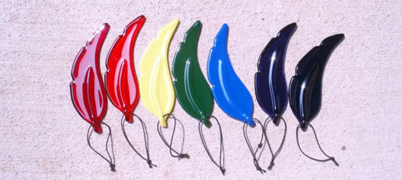 Puede incluir: Un conjunto de ocho adornos de plumas de vidrio de colores. Las plumas son de varios colores, incluyendo rojo, blanco, amarillo, verde, azul y negro. Cada pluma tiene un lazo en la parte superior para colgar.