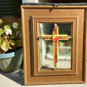 May include: A framed stained glass cross with a gold-colored wooden frame. The cross features colorful glass pieces in shades of red, yellow, green, and blue. The glass has a textured appearance, and the frame is rectangular.